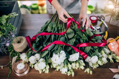 top view on young female hands tying flowers in red ribbon, preparing them for sale, wonderful beautiful white bouquet on table. floristの写真素材