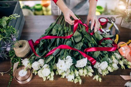 top view on young female hands tying flowers in red ribbon, preparing them for sale, wonderful beautiful white bouquet on table. florist