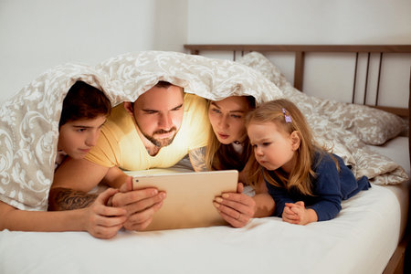 friendly caucasian family lying under blanket at home at morning, watching video on tablet, mother...