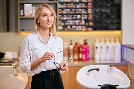 Portrait of beautiful caucasian woman consultant in white shirt, holding eye glasses in hands, selling professional shampoos and balms for beauty proceduresの写真素材