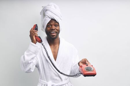 African man in bathrobe and towel talk on landline telephone isolated over white backgroundの写真素材