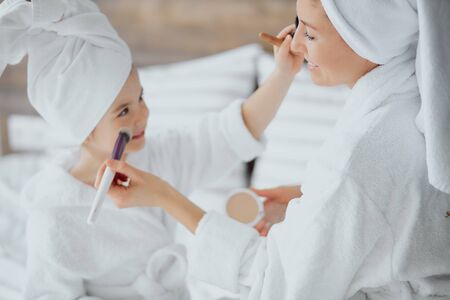 caucasian woman and little daughter sit on bed wearing towels and bathrobe doing makeup after shower. family conceptの写真素材