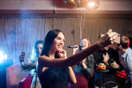handsome guys and beautiful ladies in dressed have party time in karaoke, modern caucasian people singing in microphoneの写真素材
