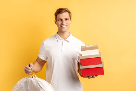 portrait of young caucasian courier man with boxes and clothes for customer isolated over yellow...