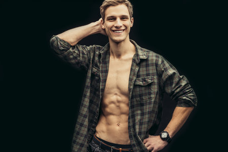portrait of beautiful caucasian young man smiling at camera, male with perfect athletic body look at camera, wearing casual checkered shirt isolated over black backgroundの写真素材