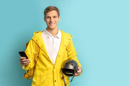 portrait of good-looking deliverman in special yellow uniform, using smartphone, helmet from motorcycle and carrying orders for customers isolated over blue backgroundの写真素材
