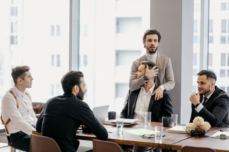 funny caucasian business colleagues sit on table and have conversation during free time at work, everyone in formal wear. one man grabbed the head of another. panoramic window in the backgroundの写真素材