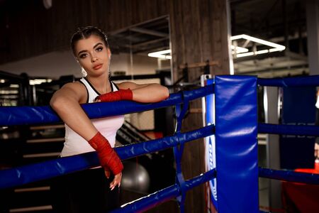 Tired woman boxer after intense training in ring, stand leaning on fence, effective training in gym.の写真素材