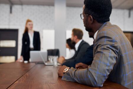Young caucasian lady training young international group of business people, discussing strategy of building new business. African man in tuxedo and eyeglasses, side viewの写真素材