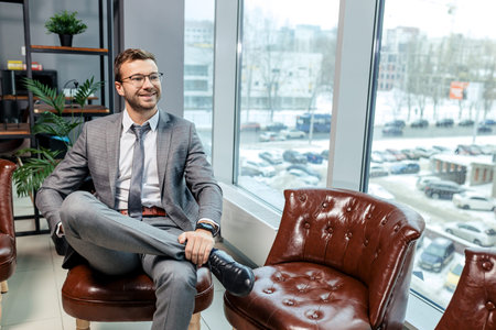 young caucasian business male in elegant stylish suit sit on leather chair in executive boardroom after business meeting with partners, personable man with beard sit near panoramic windowの写真素材
