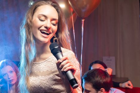 young emotional female singing in microphone in karaoke bar, wearing dress, friends with balloons celebrating in the backgroundの写真素材