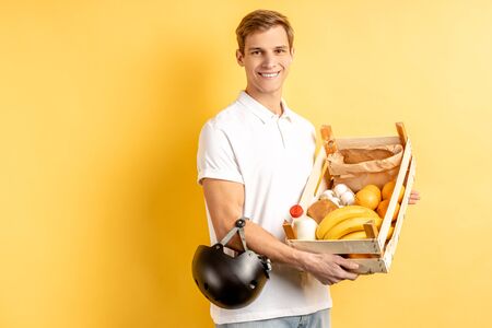 portrait of smiling young delivery man holding fresh vegetables and fruits in box, look at camera. isolated over yellow backgroundの写真素材