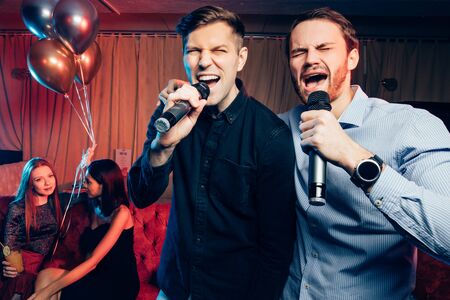 young caucasian friends men in shirts singing in microphone in karaoke bar, having fun, celebrating. holiday, leisure, party conceptの写真素材