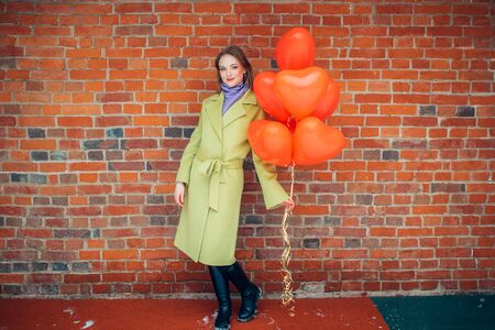 portrait of young and beautiful caucasian lady in coat with red air balloons. good-looking woman 20-25 yeard ols stand posing and looking at camera. red brickwall in the backgroundの写真素材