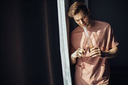 young caucasian man lighting bong with cannabis at home, indoors. guy stand next to window. unhealthy lifestyle, cannabis, marijuana, smoking issues, hemp medicine conceptの写真素材