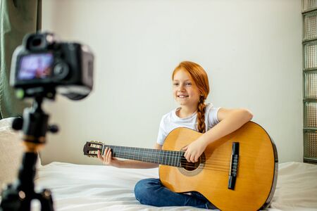 young blogger perform music at camera, red haired little girl play acoustic guitar, self-taught girl present her new song for subscribersの写真素材