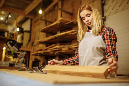 young caucasian professional carpenter woman checking for roughness piece of wood after grinding, female in uniform work in factoryの写真素材