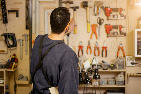 rear view on young carpenter in uniform taking hammer for making wooden furniture, in factory