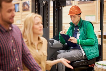 young man in uniform and helmet, warehouse worker compares product data, reads a document and takes notes while customers pass by him in the marketの写真素材