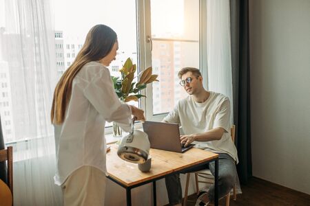 kind caucasian woman treats her husband with tasty tea while he is working from home, using laptop. merciful wife loves husband, support him. freelance, relationships, business at homeの写真素材