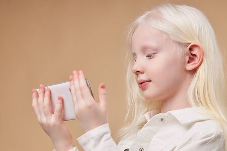 beautiful albino child holding white smartphone in hands isolated, girl with white hair and skin look at screen on mobile phone. unusual beauty of people and digital modern technologies conceptの写真素材