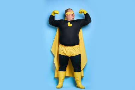 handsome brave strong man has a mad desire, a dream to shield the world from dirt and evil. fat superhero in yellow costume posing isolated over blue backgroundの写真素材