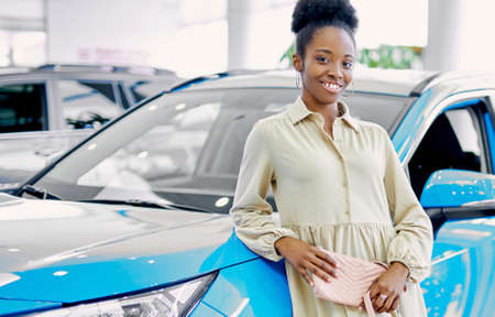 portrait of young slim african woman standing next to new blue car, attractive lady in dress posing at camera, make purchase in dealershipの写真素材