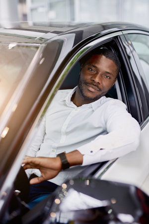 young black man happy with purchase in dealership, he is owner of represntative modern car, male...