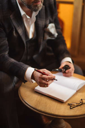 Caucasian middle-age professional businessman putting notes in paper organiser sitting on armchair in restaurant.の写真素材