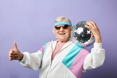 Portrait of happy senior woman in white sportswear holding discoball in hands, posing isolated over purple backgroundの写真素材