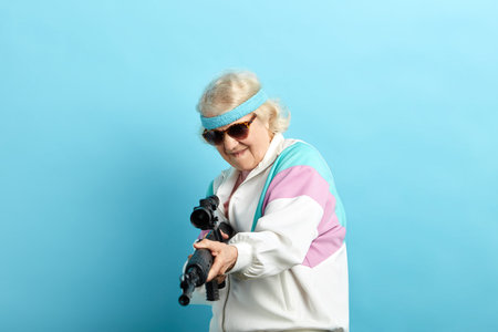 Senior woman in sports wear and black sanglasses screaming while aiming at camera with a rifle...