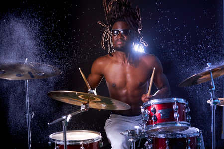 wet drum set covered with water drops, african young man beating on drum set with sticks. skillful black drummer with naked skin and dreadlocksの写真素材