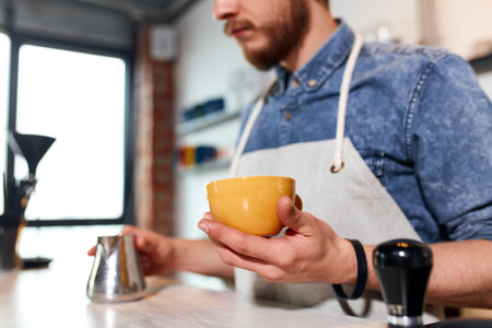Skillful bearded man takes small metal milk jug from light coloured wooden counter, holds small size...