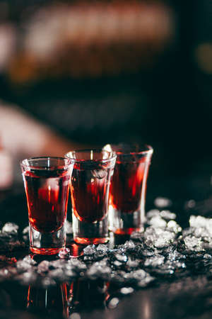 Three red alcohol shots standing in line surrounded with ice pieces, isolated on dark blurred background. Concept of alcohol, spirits for a good night time.の写真素材