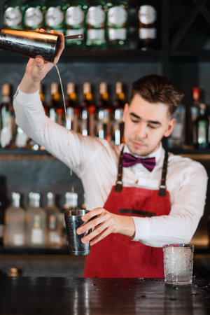 Creative barman making alcoholic drink, pouring fresh cocktail from shaker into matalic glass on the...