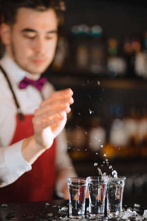 Bartender in the white shirt and red apron adding ice crumbs into an empty cocktail glass on the bar counter at restaurantの写真素材