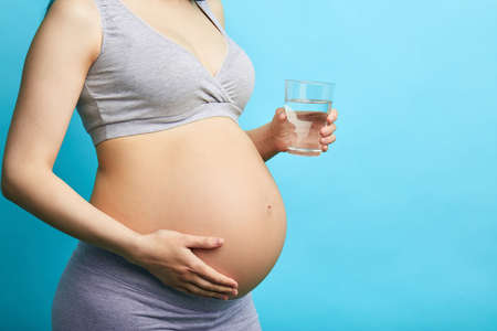Young pregnant caucasian model on third trimester, dressed in grey underwear, holding a glass of water, isolated on light blue baclground with copyspace.の写真素材