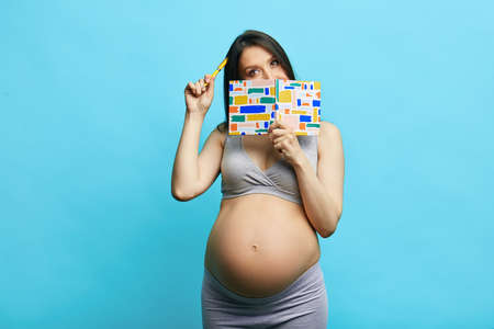 Pregnant young woman with brown long hair, standing isolated in underwear writing in notebook her notes about her health condition standing against blue wallの写真素材