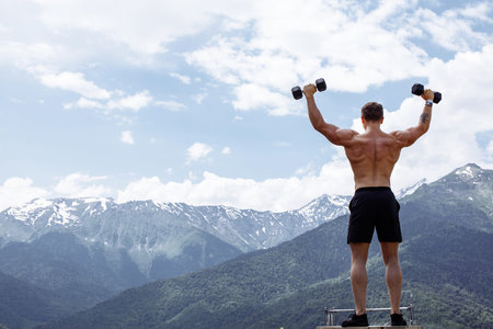 Young and sporty muscular half naked crossfit male athlete training with dumbbells outdoor over...
