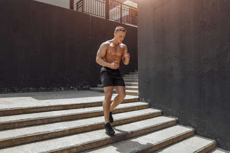 Blonde european male fitness model with naked torso, wears black sportive shorts, running on staircase, goes in for sport regularly. People, outdoor sports and exercising conceptの写真素材