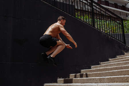 Handsome strong man doing warming up plyometric exercise in the mid-air. Young caucasian man with naked torso jumping upstairs while exercising outsideの写真素材