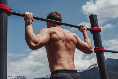 Portrait of a motivated shirtless sportsman doing pull-ups exercise on horizontal bar outdoors, over...