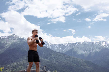 Young and sporty muscular half naked crossfit male athlete training with dumbbells outdoor over green mountains and blue sky background. Sport, health, outdoor athletics.の写真素材