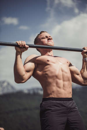 Caucasian male athlete with fit torso and strong hands doing chin ups outdoor. Handsome blonde sportsman pulls up on stadium horizontal bar. Chin up bar exercises.の写真素材