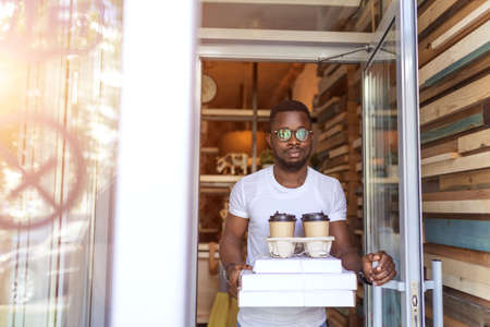 Here is your order, sir. African american delivery guy standing at house porch, holding a stack of boxes with freshly made pizza and take away coffee.の写真素材