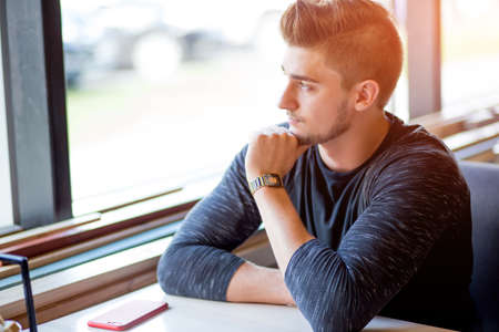 Young handsome bearded caucasian man sitting near the window, looking with pensive expression at the window at a seaside restaurant. Waiting for his coffee.の写真素材