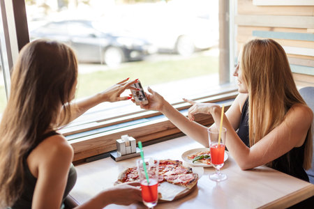 Young women, blonde and brunette meeting at restaurant after a long break, eating pizza and sharing...