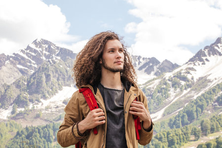 Young curly haired Man Traveler with red backpack relaxing outdoor with rocky mountains on...