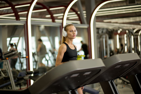 beautiful blond woman runs in gym on treadmill with big white headphones, girl listens to music,...