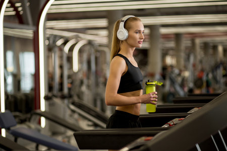 blond woman Running in sports club on treadmill with water bottle and listen music with white...
